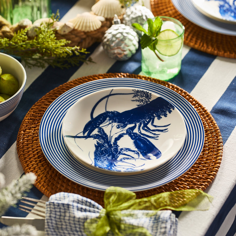 A table setting featuring the Caskata Lobster Coupe Salad Plate on a striped plate and wicker charger, paired with a green drink, lime, and decorative accents atop a blue and white striped tablecloth.