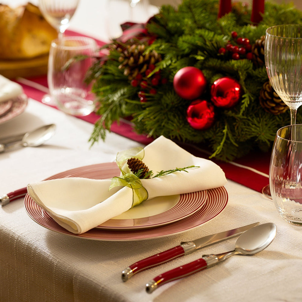A festive holiday table features Caskata’s Newport Crimson Stripe Table for 4, accented by red-handled cutlery, a pinecone napkin, and a centerpiece of greenery and red ornaments.