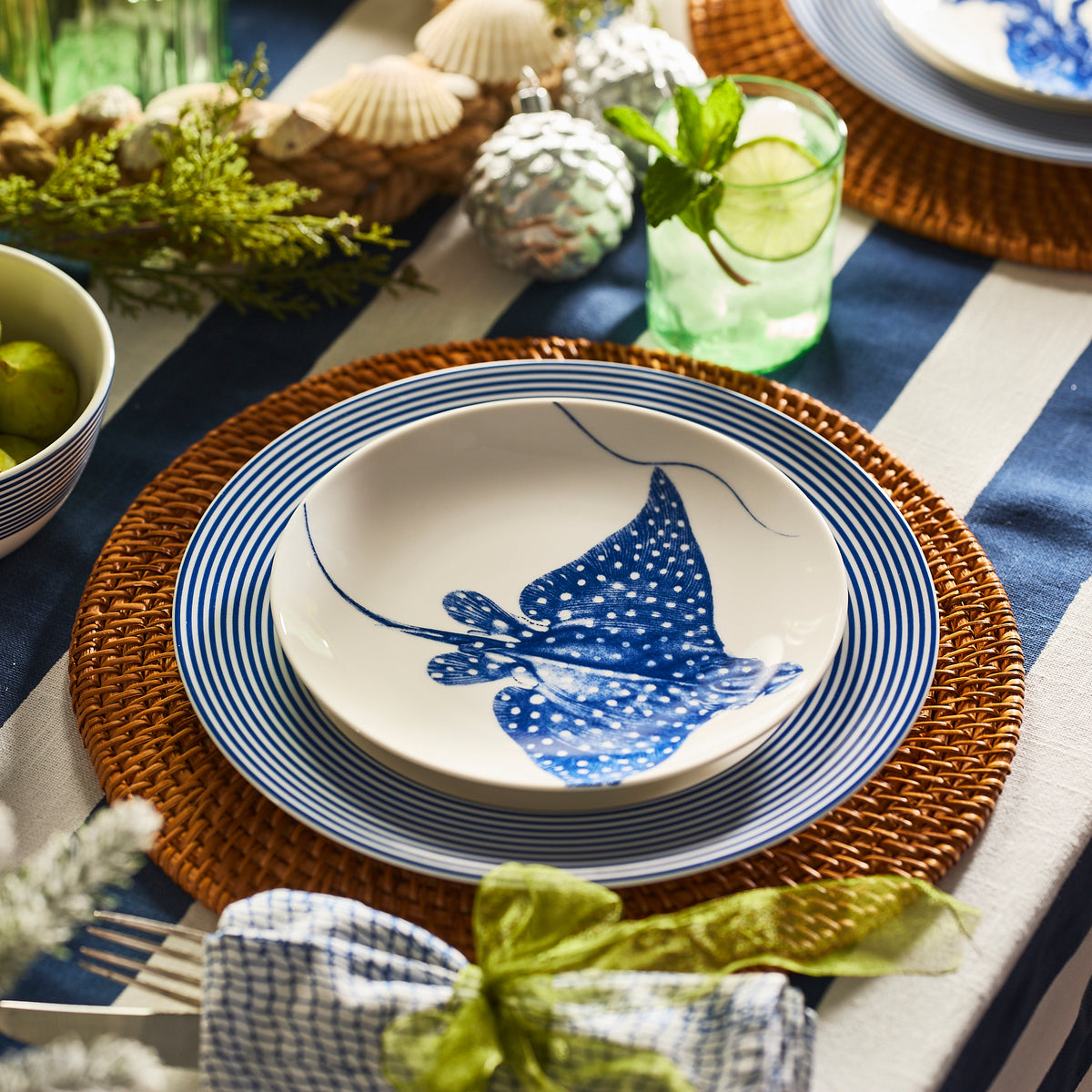 A close-up of a table set with Caskata&#39;s Stingray Coupe Salad Plate, striped blue and white dinnerware, a wicker placemat, and a glass garnished with lime and mint.