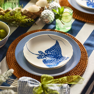 A close-up of a table set with Caskata's Stingray Coupe Salad Plate, striped blue and white dinnerware, a wicker placemat, and a glass garnished with lime and mint.