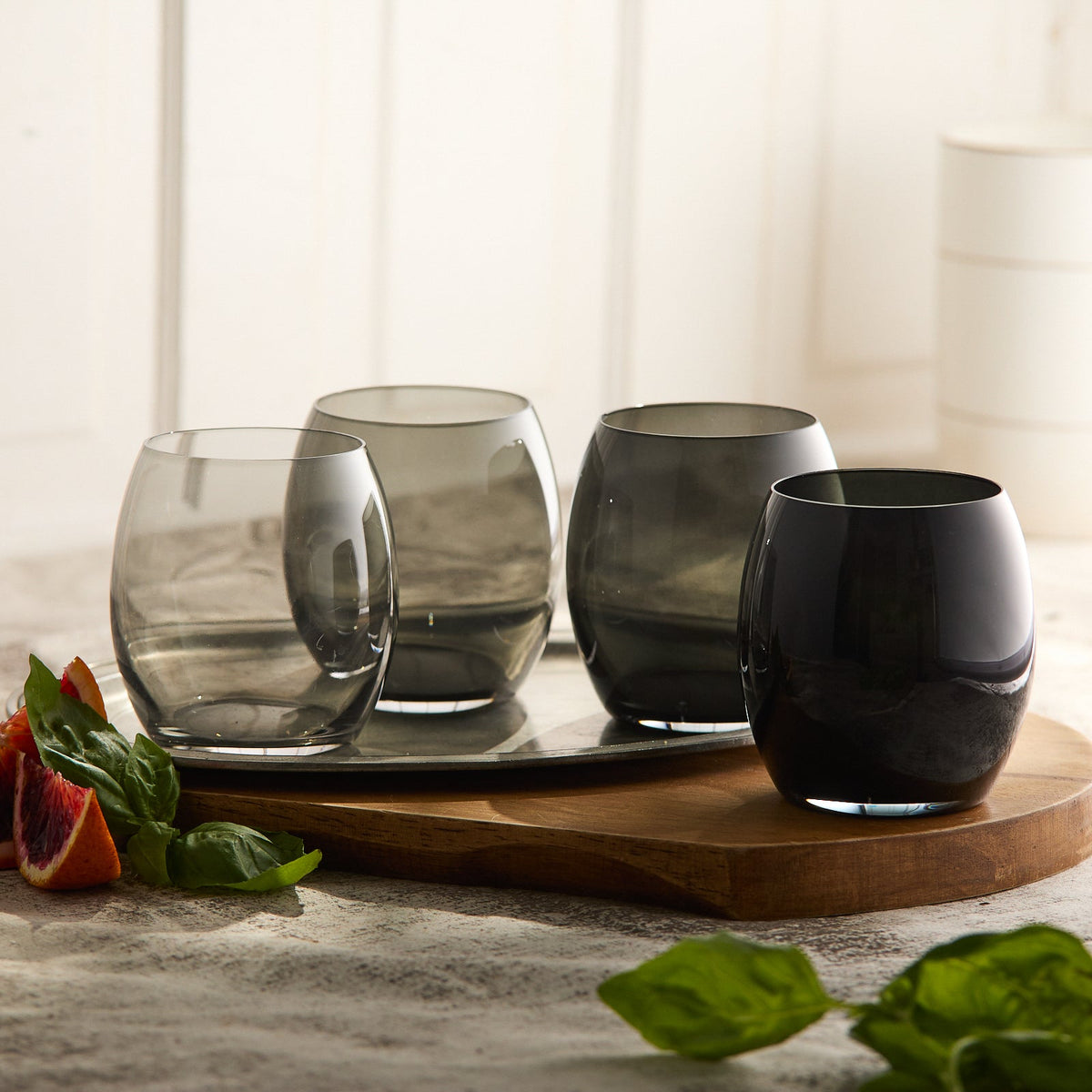 The Les Nuages Gray Ombré Glasses Set of 4 by Caskata features lead-free crystal tumblers in clear to dark gray, artfully placed on a wooden board with leafy greens nearby, crafted meticulously in Slovakia.