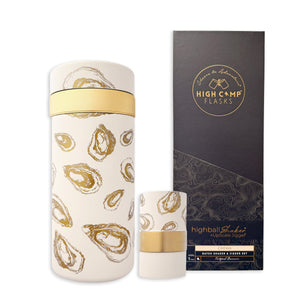 The Caskata x High Camp Oysters Batch Cocktail Shaker by Caskata—a white, vacuum-insulated shaker with gold oyster designs—sits beside its chic black-and-gold box, making a stylish bar accessory.