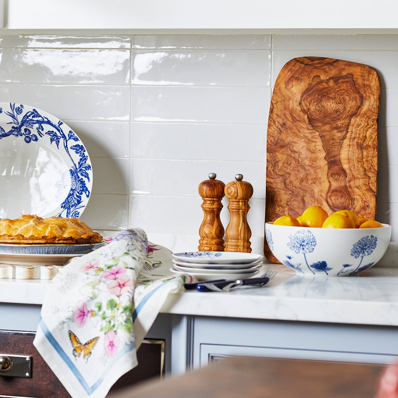 A kitchen counter displays a delectable pie, floral dinnerware plates, a decorative towel, pepper mills, a wooden board beside Caskata Artisanal Home's Summer Blues Vegetable Serving Bowl of lemons against the white tiled backsplash.