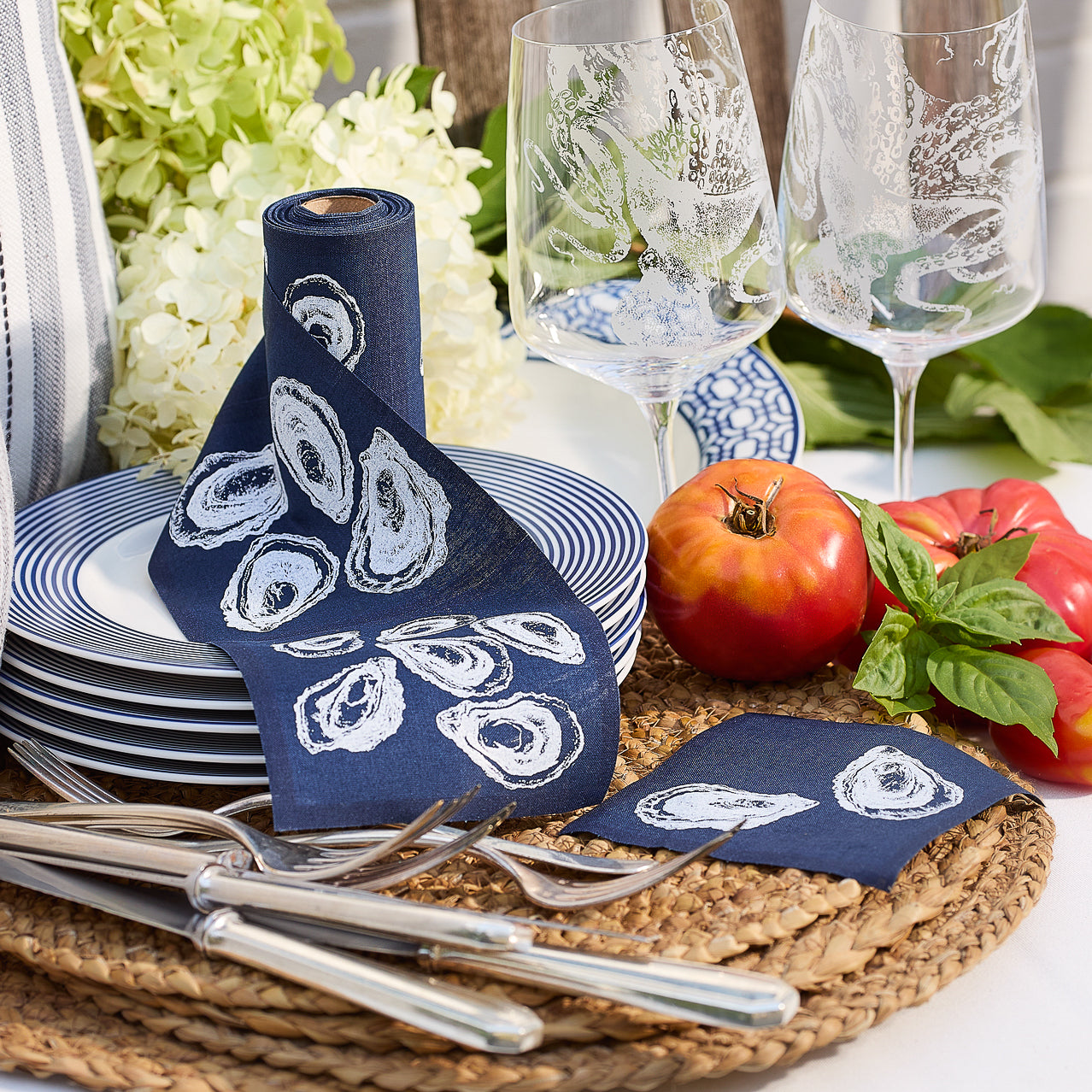 An Oysters Cocktail Napkin Roll by MY DRAP in dark blue with white oyster shell prints, paired with two coordinating square sheets, styled gracefully on a beige surface.