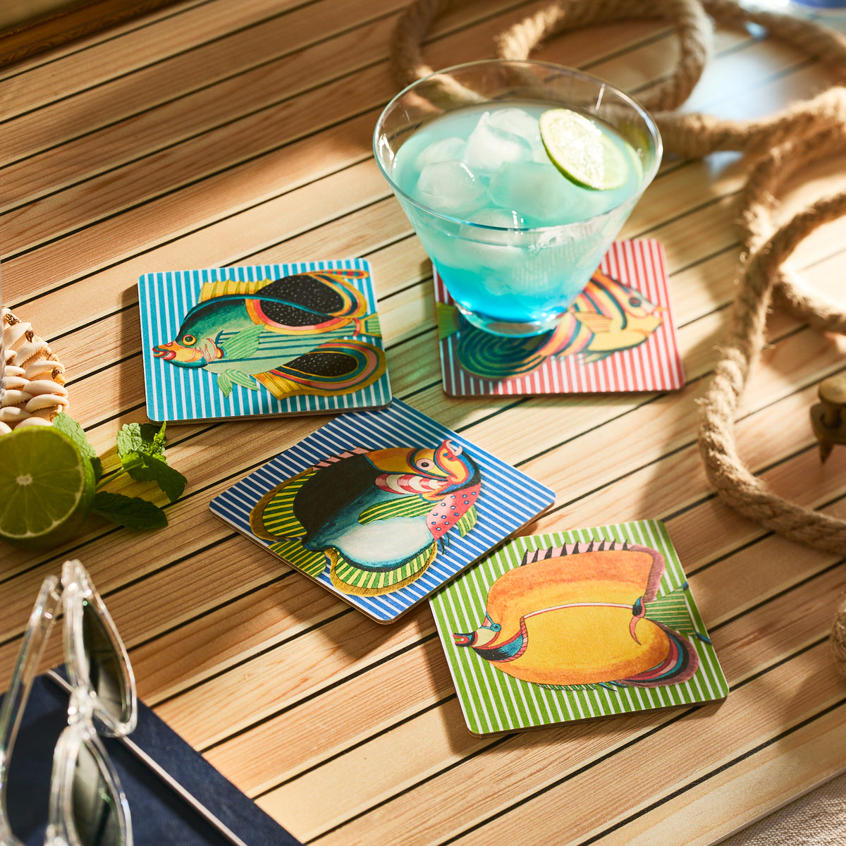 A blue cocktail with ice and lime sits on an FSC-Certified coaster, surrounded by Caskata&#39;s Party Fish Birchwood Coasters, Set of 4, on a wooden table. Nearby birchwood trays hold glasses, a lime, mint, and rope for a sustainable touch.
