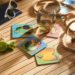 A wooden table adorned with Caskata’s Party Fish Birchwood Coasters, a glass of white wine, sunglasses, a book, rope, lime, and seashells. FSC-certified birchwood trays nearby enhance the eco-friendly charm of this seaside vignette.