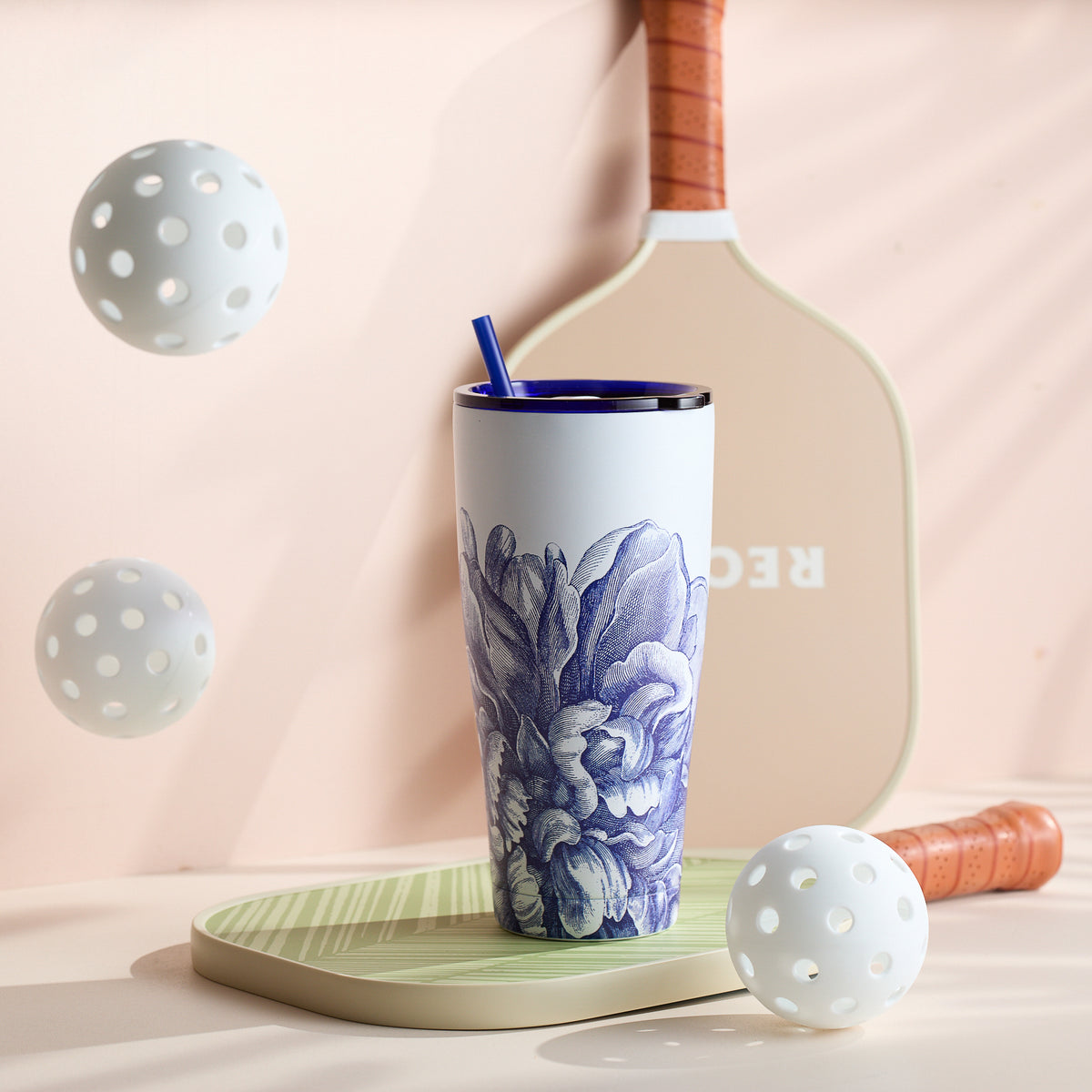 A Caskata Peony Stainless Steel Insulated Tumbler with a blue straw sits on a pickleball paddle, flanked by two perforated balls.