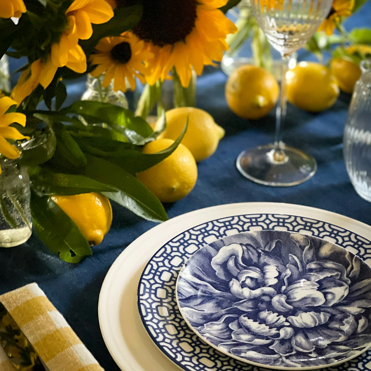 A table setting with heirloom-quality blue and white Peony Small Plates by Caskata Artisanal Home featuring floral-patterned plates, a yellow striped napkin, lemons, and a bouquet of yellow flowers in the background.