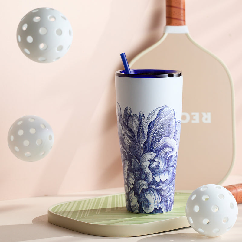 The Caskata Peony Stainless Steel Insulated Tumbler, featuring a blue straw, rests on a green surface alongside a pickleball paddle and two white pickleballs. It keeps your drink perfectly tempered.
