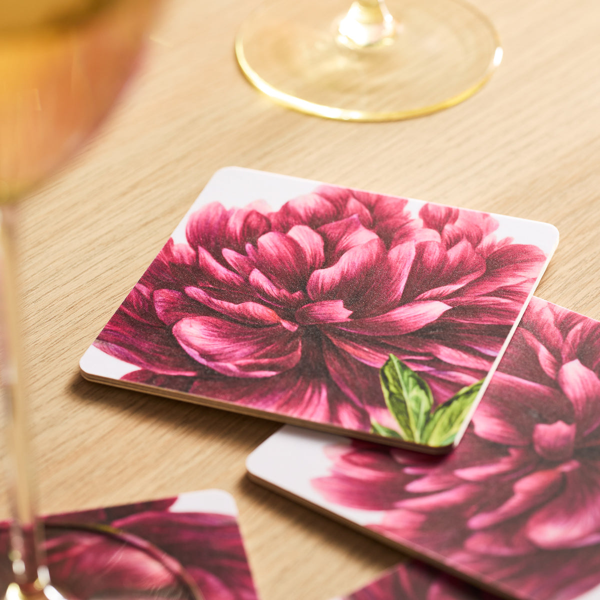 Pink Peony Birchwood Coasters by Caskata, adorned with red florals, rest on a wooden table near a partially visible glass on FSC-Certified Birchwood trays.