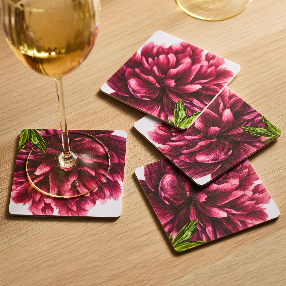 A wine glass sits elegantly on a Caskata Pink Peony Birchwood Coaster, set of 4, with FSC-certified trays scattered across the wooden table.