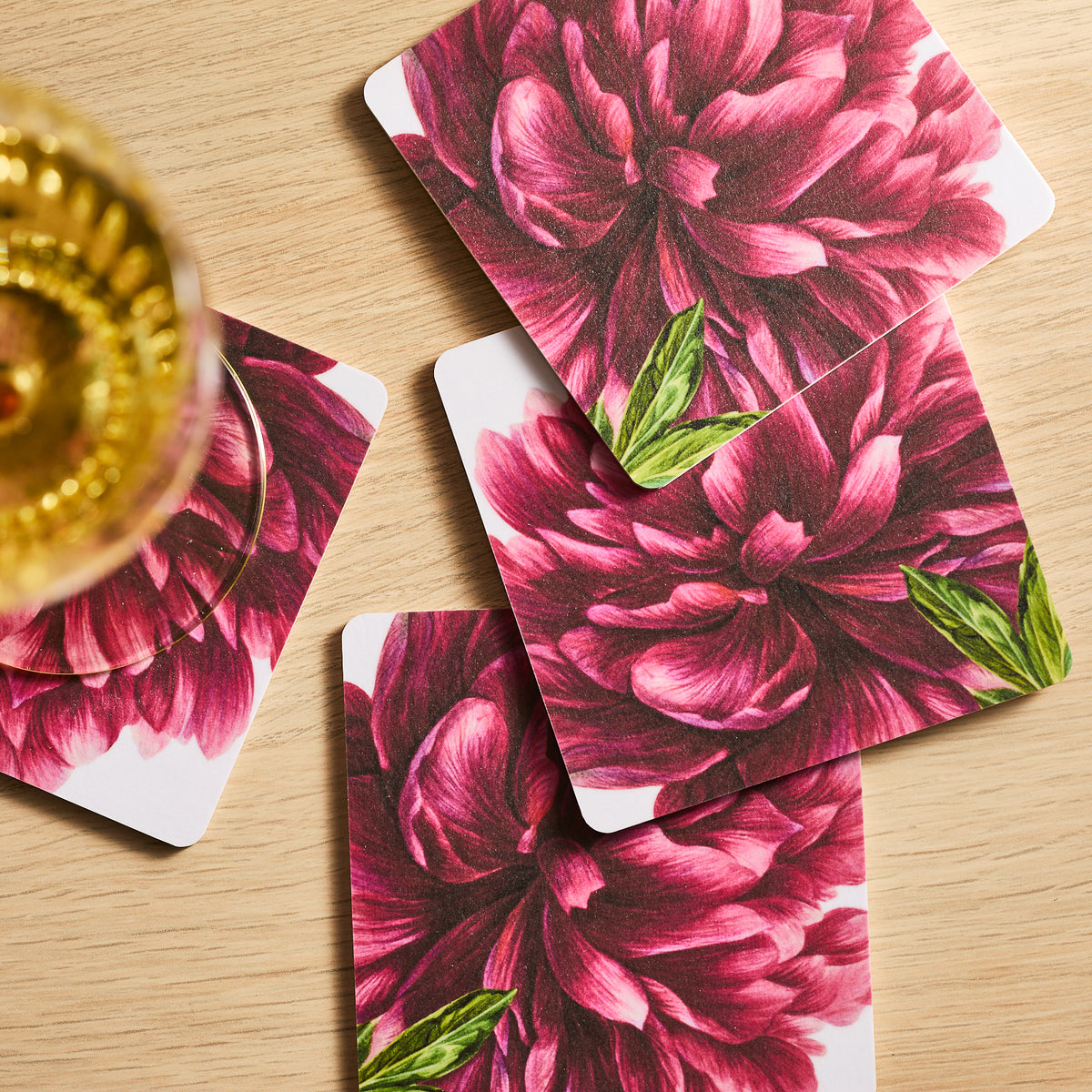 The Pink Peony Birchwood Coasters, a set of four by Caskata, feature red floral designs with green leaves on FSC-Certified Birchwood. Their natural tones complement the light wood surface, while a wine glass nearby adds elegance to this eco-friendly setting.