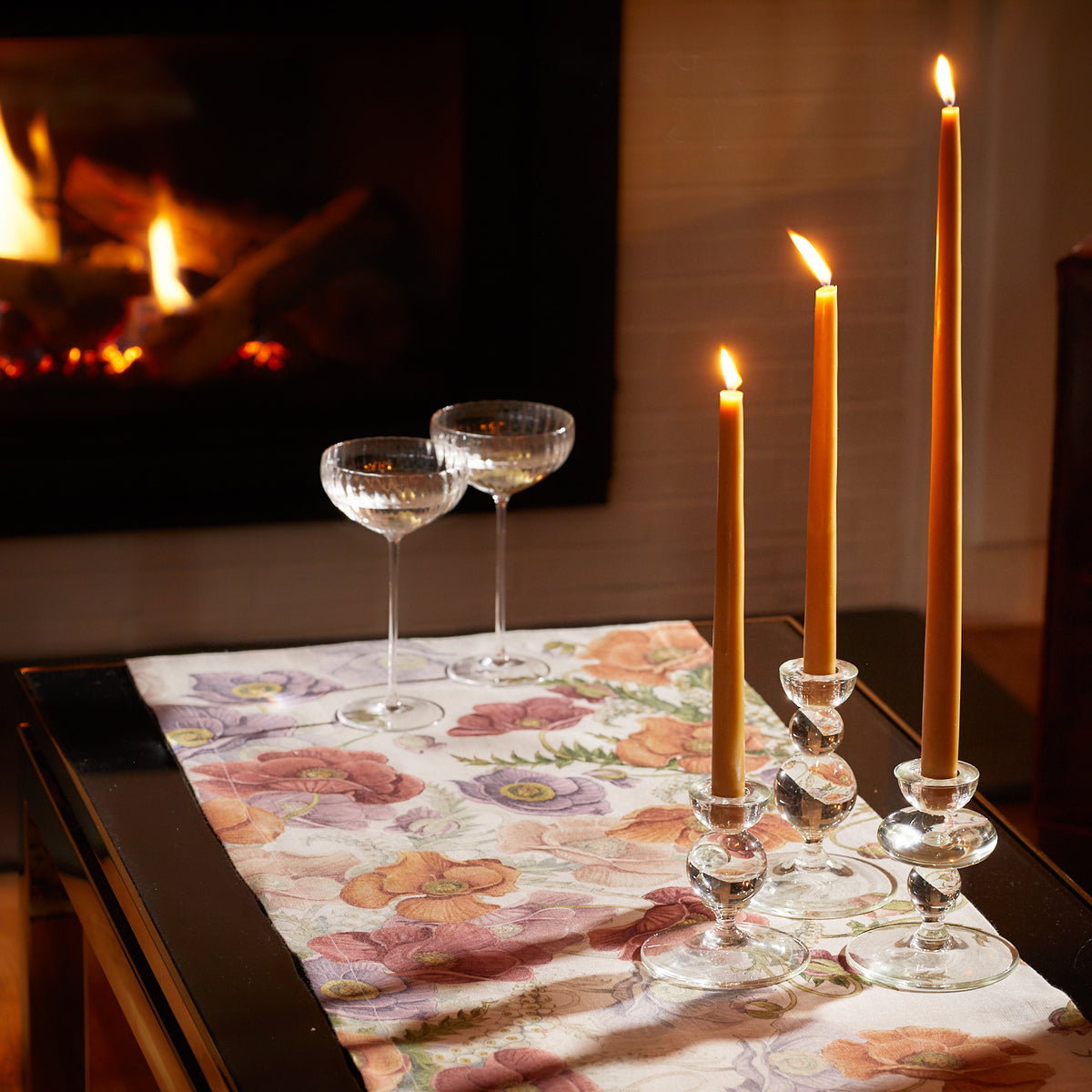 A table with Caskata X The Pool Balance Glass Candlestick Holders Set/3 and wine glasses in front of a fireplace.