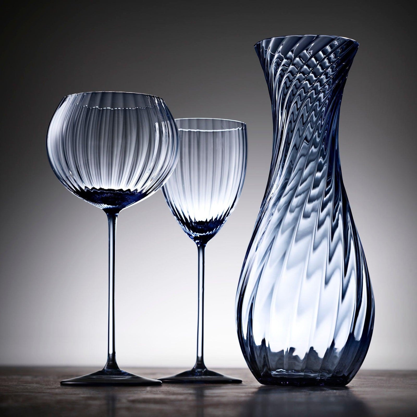 The Quinn Ocean Wine Lovers Set by Caskata features a blue-tinted glass carafe, four goblets, and eight crystal wine glasses with matching swirl patterns and stems, arranged on a white background—the ideal set for wine lovers.