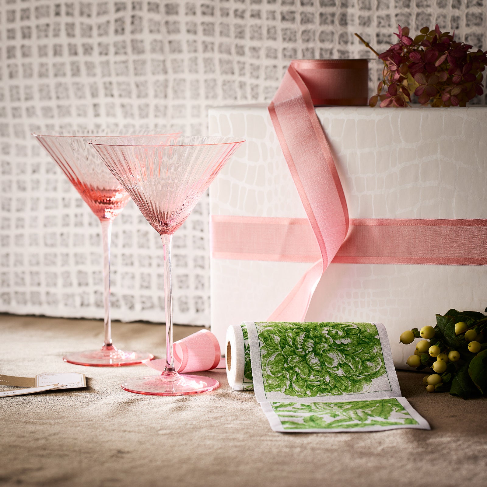 The "Cheers to You" Bundle by Caskata features two crystal martini glasses, a gift wrapped with a pink ribbon, green floral fabric, and a small bouquet arranged beautifully on a textured surface.