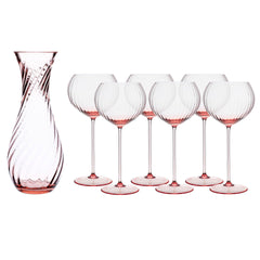 The Quinn Rose Red Wine Lover Set by Caskata includes six handmade wine glasses with pink-tinted stems and bases, plus a matching pink-tinted carafe featuring a swirled pattern—perfect for stylish gatherings.