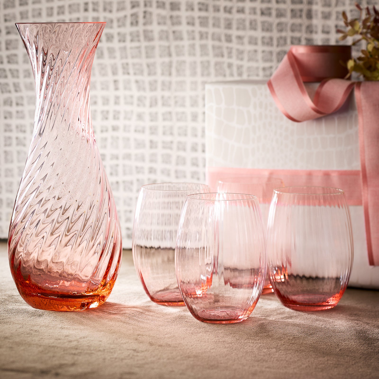 The "Sweet Life" Bundle by Caskata features a textured rose pink carafe and three matching crystal stemless wine tumblers, shown on a table with a white gift box tied with a pink ribbon in the background.