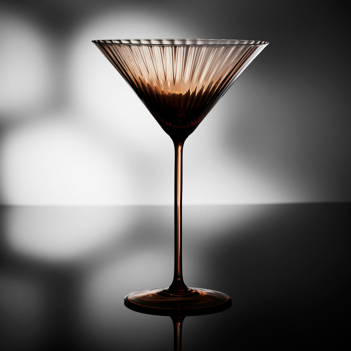 Quinn Amber Brown Oversized Martini Glasses Set of 2 in mouth blown crystal from Caskata, featured on a backlit moody tabletop.