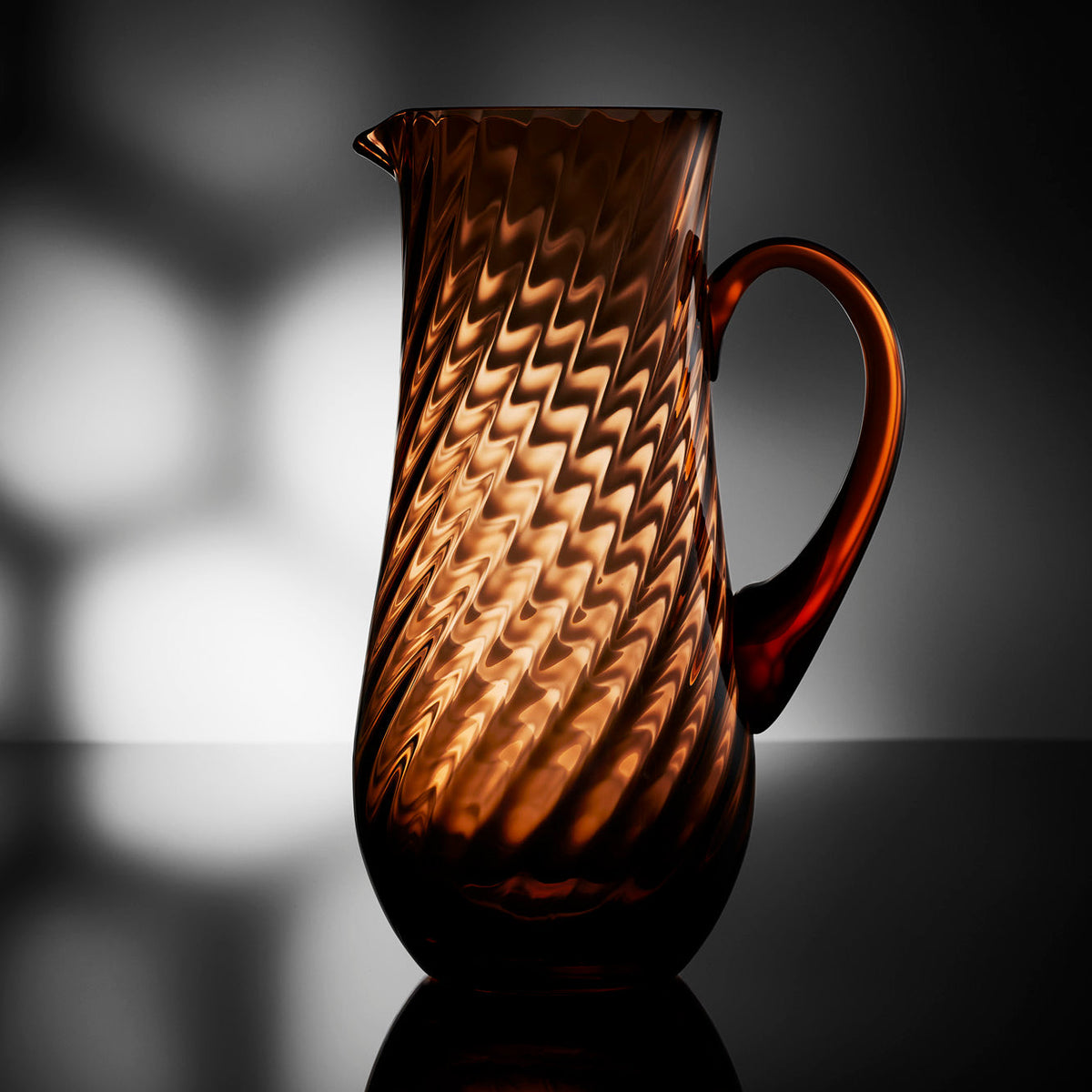 Quinn Amber Pitcher in mouth blown crystal from Caskata, on a moody backlit tabletop.