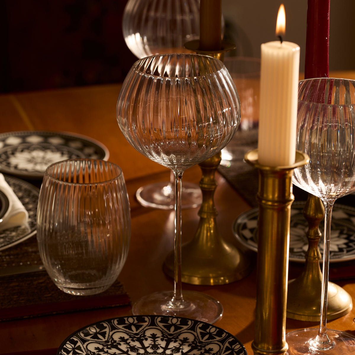 Elegant dining table set with patterned plates and Quinn Clear Red Wine Glasses by Caskata Artisanal Home, alongside lit candles in gold holders.