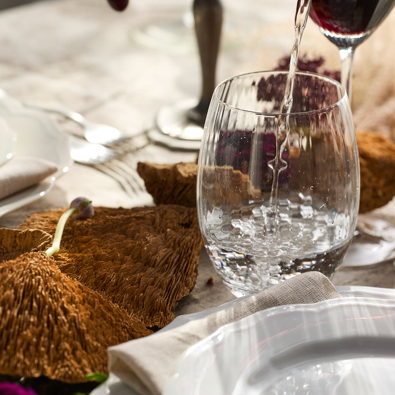 Water cascades into a Quinn Clear Tumbler, a piece from Caskata Artisanal Home, resting elegantly on a table with a plate, napkins, and charming brown decor. Crafted from lead-free crystal in the Czech Republic, this setting embodies sophistication and craftsmanship.