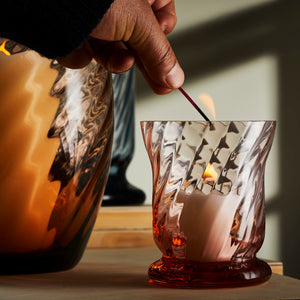 A hand lights a candle inside a red-tinted, textured glass holder from the Quinn Rose Candlelight Bundle by Caskata, placed on a wooden surface next to a large, mouth-blown Czech Republic wavy glass vase.