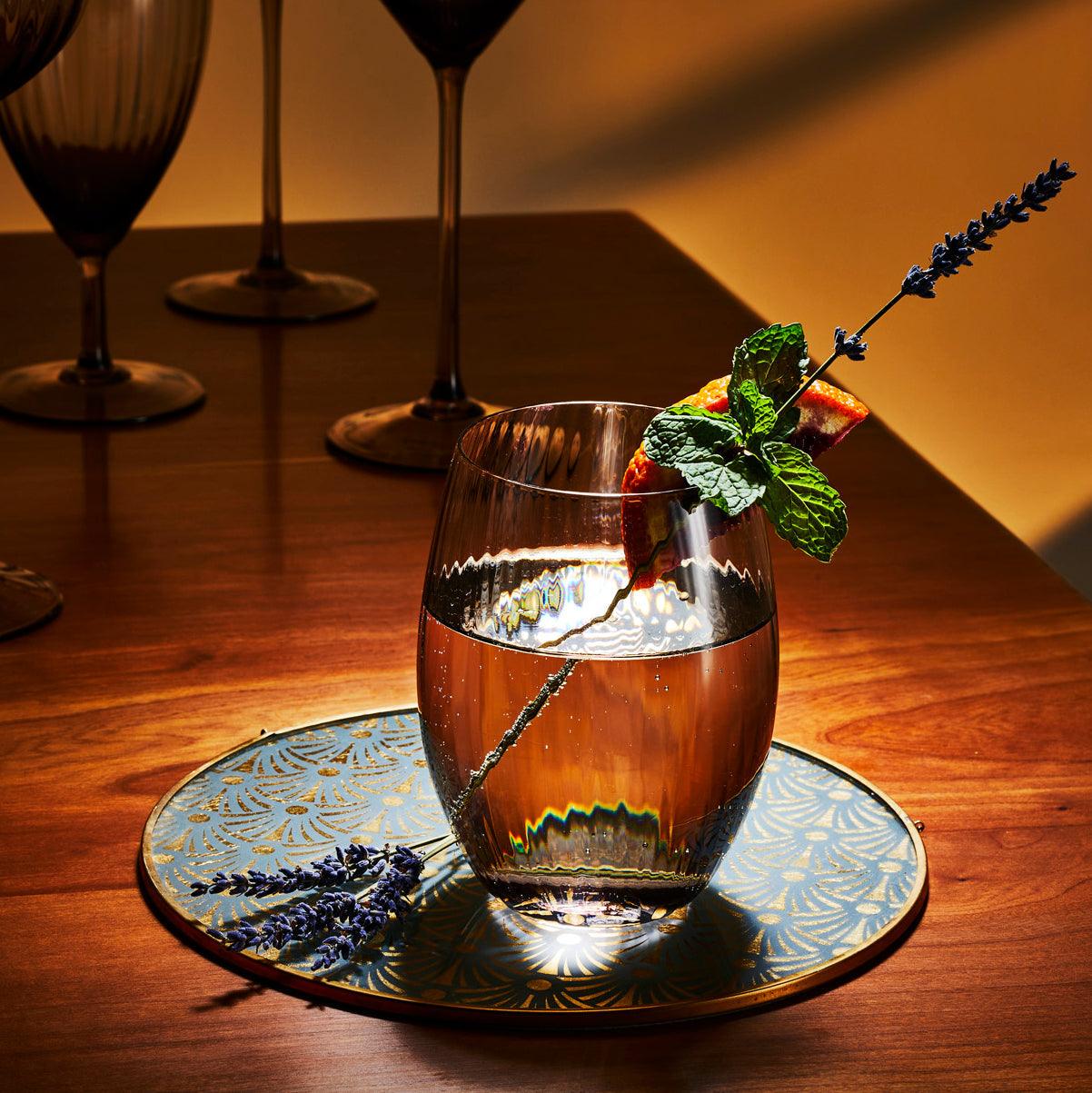 The Quinn Mocha Dinner Party Set by Caskata features a textured glass carafe and six ribbed crystal glasses in a light brown tint, creating an elegant display on a white background.