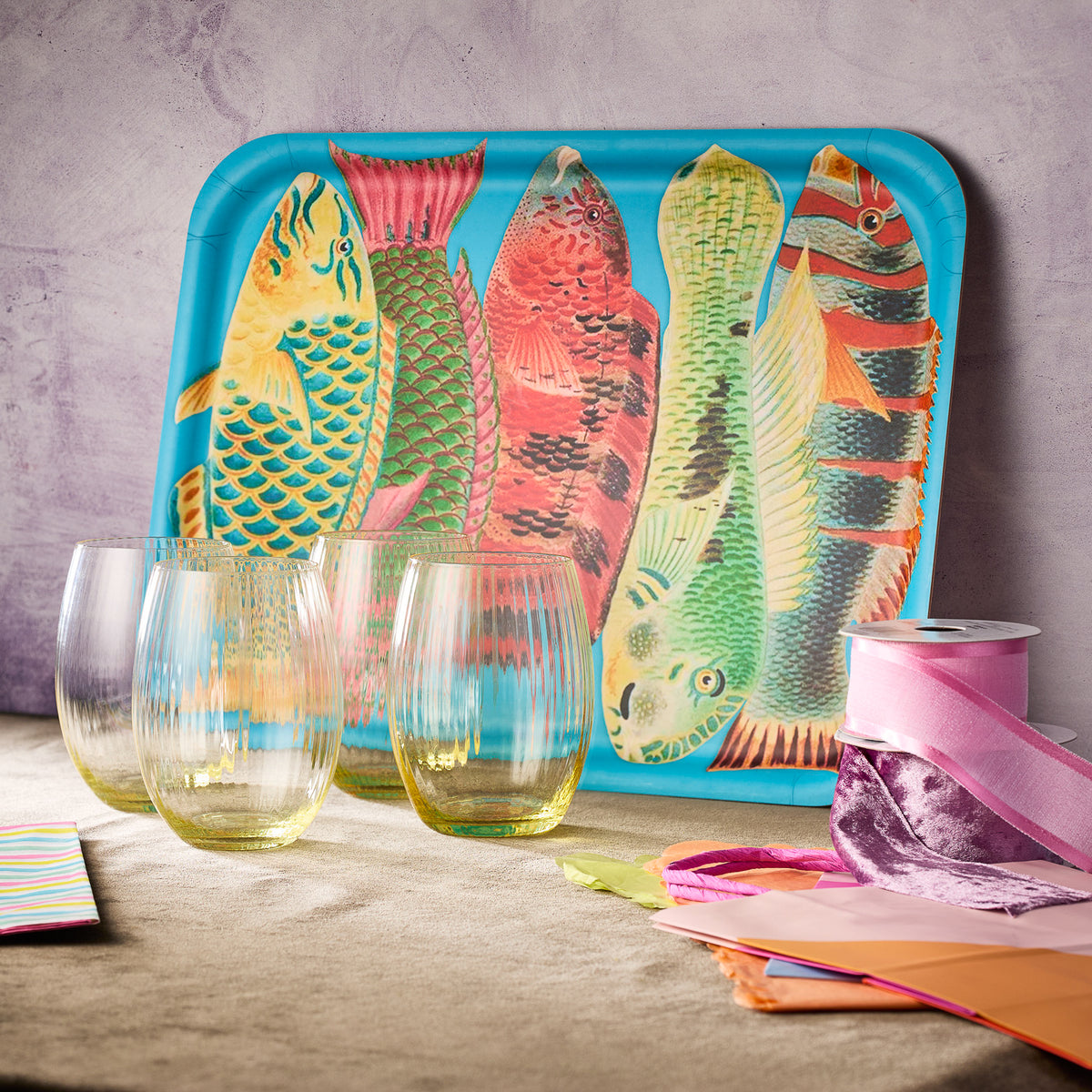Caskata's "Tropic Fun & Sun" Bundle features a birchwood tray with bright fish artwork, four ribbed stemless wine glasses, and pastel ribbons and paper displayed on a beige surface.