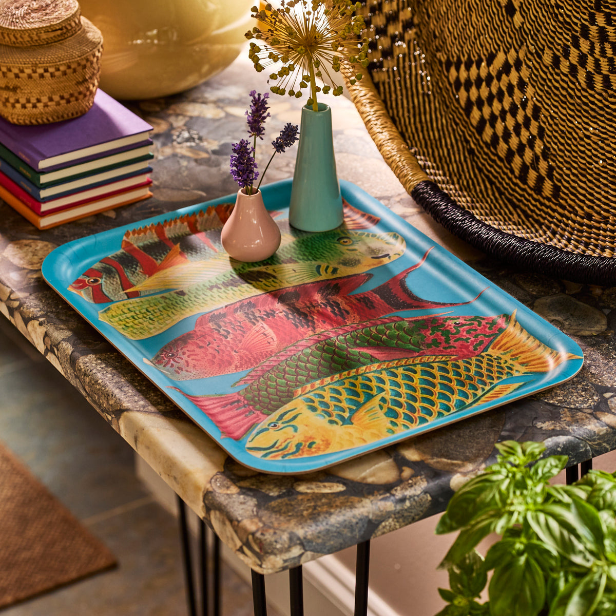 A table with Caskata&#39;s Rainbow Fish Large Rectangular Tray, a blue sustainable piece featuring colorful fish, alongside small flower vases, stacked books, woven baskets, and a green plant in the foreground.