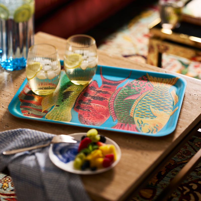 A Caskata Rainbow Fish Large Rectangular Tray holds two drinks with lemon and ice on a wooden table, next to a small plate of fruit and a fork on a napkin.