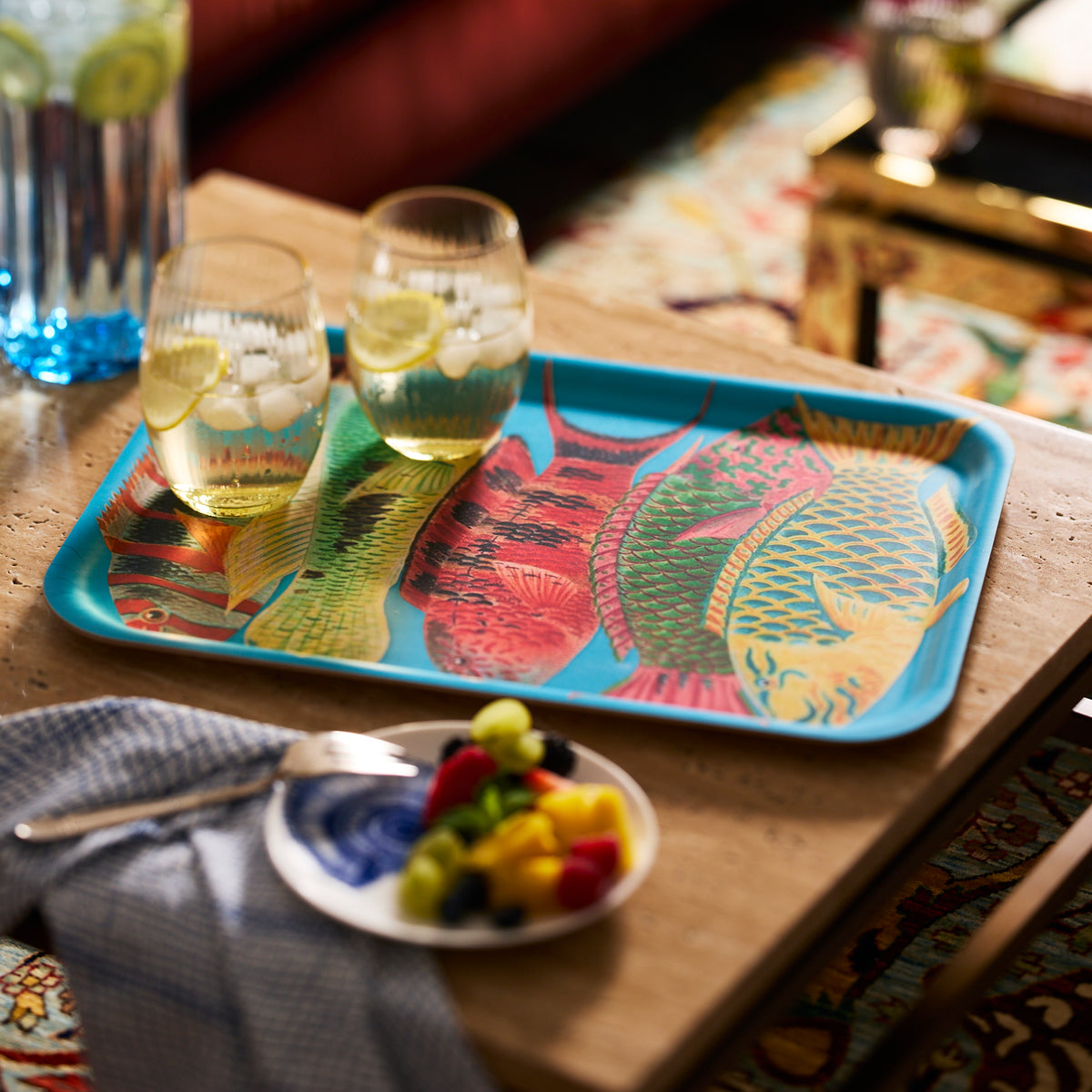 A Caskata Rainbow Fish Large Rectangular Tray holds two drinks with lemon and ice on a wooden table, next to a small plate of fruit and a fork on a napkin.