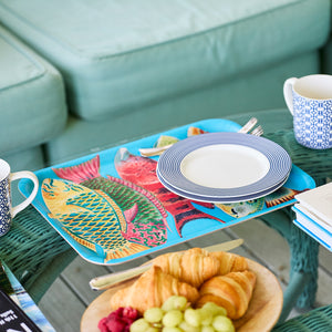 The Caskata Rainbow Fish Large Rectangular Tray holds a plate and cutlery on a glass table. Nearby are patterned mugs, a plate with croissants and fruit, plus books.
