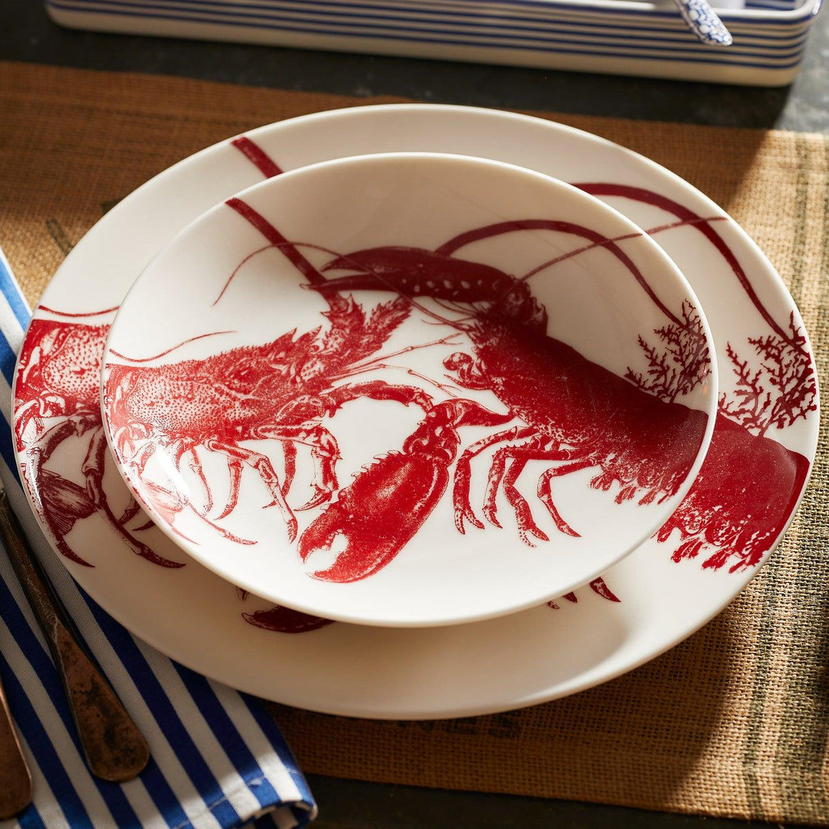 A close-up of white dishware featuring red lobster illustrations. A large plate and a Caskata Lobster Coupe Salad Plate with similar designs are stacked on a woven placemat next to a blue and white striped napkin, enhancing the seaside style.