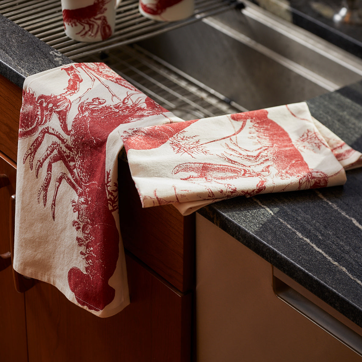 The Lobster Love Bundle by Caskata showcases two kitchen towels with red lobster and crustacean prints elegantly draped over a dark countertop near a sink.