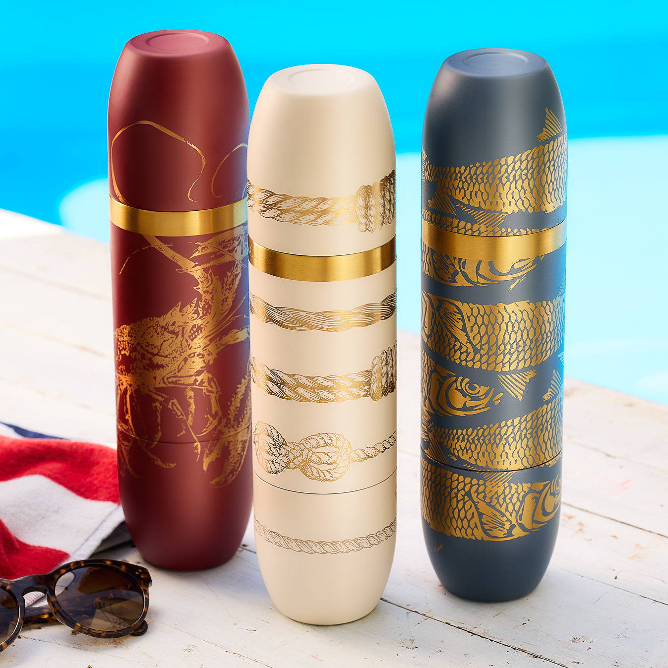 Caskata’s Summer Adventures Flask Bundle features three insulated wine bottles—lobster on red, knots on white, fish on blue—displayed by a pool with sunglasses and a towel for the perfect stylish summer set.