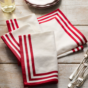 Two Triple Stripe Red Dinner Napkins by Caskata are folded on a wooden table beside stacked plates, a glass bottle, and metal cutlery. These stylish cotton napkins are machine washable for easy care.
