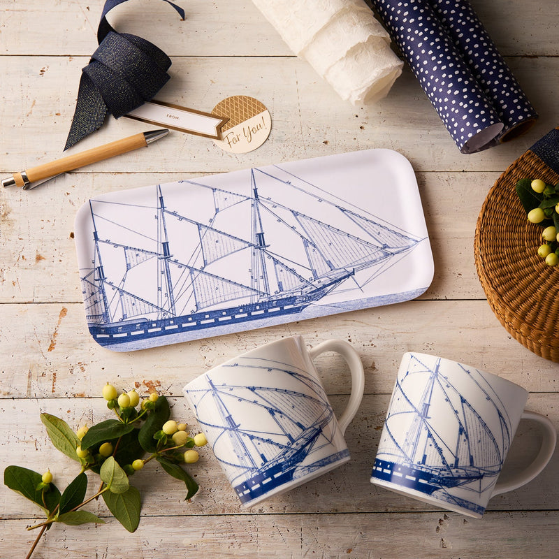 The "Clipper Cuppa" Bundle by Caskata features a rectangular decorative tray and two oversized porcelain mugs with blue ship designs—a perfect maritime gift set, beautifully arranged with gift wrap, a pen, tag, greenery, and a wicker basket.