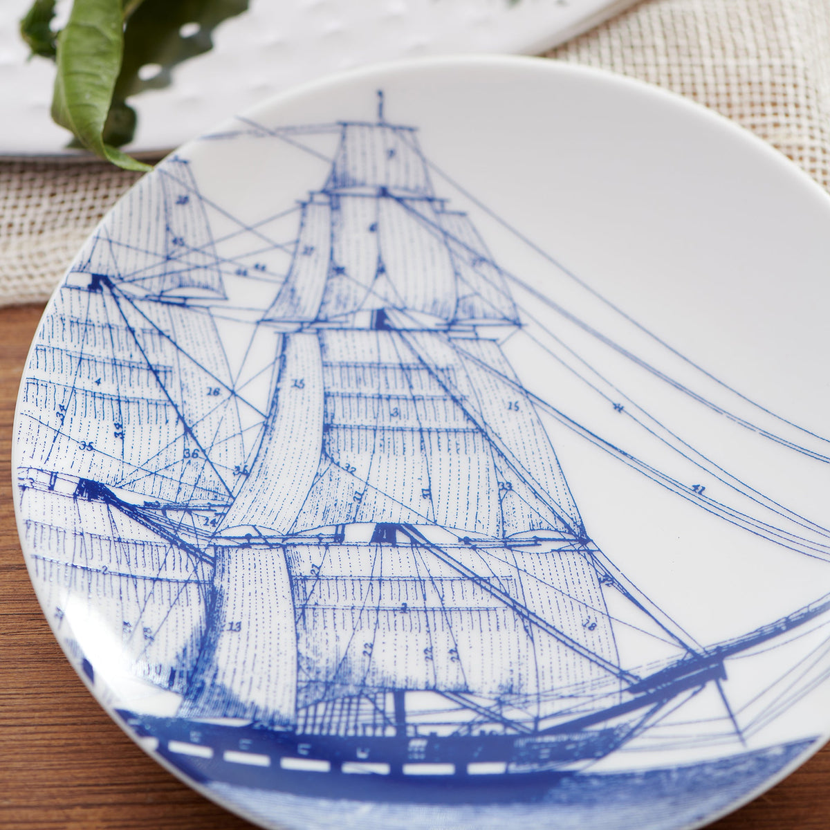 A white ceramic plate from Caskata Artisanal Home showcases a detailed blue illustration of a sailing ship with rigging and sails, reflecting nautical heritage. Set on a wooden surface with green leaves partially visible in the background, this piece embodies heirloom-quality dinnerware called Rigging Small Plates.