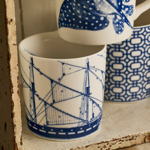 Three Caskata Rigging mugs with coastal blue designs sit on a rustic shelf: one features a ship, another displays a geometric pattern, and the third shows part of a sea creature illustration.
