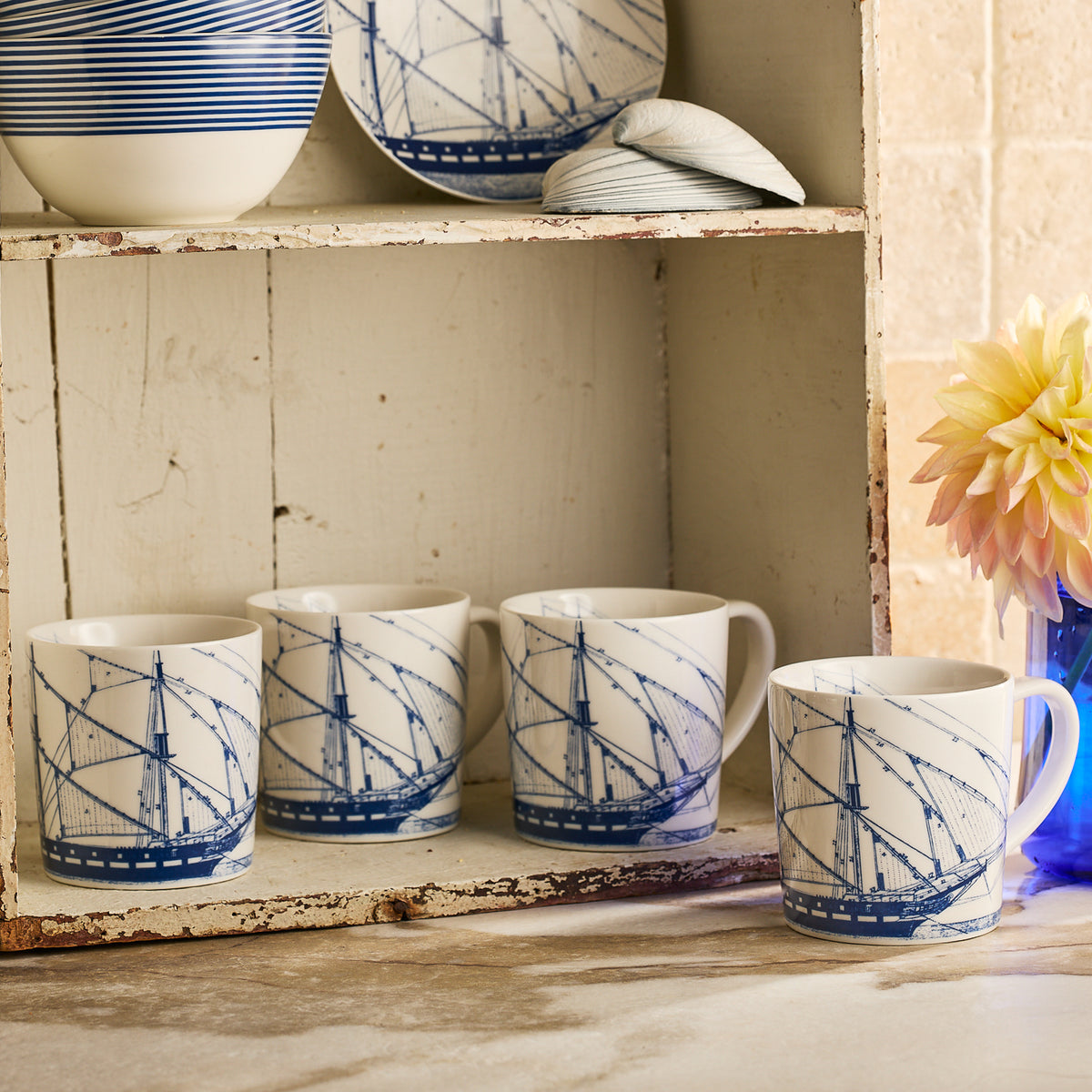 Four Caskata Rigging Mugs, featuring blue ship designs, are lined up on a rustic white shelf with blue and white ceramics and a flower vase, creating a charming coastal display.