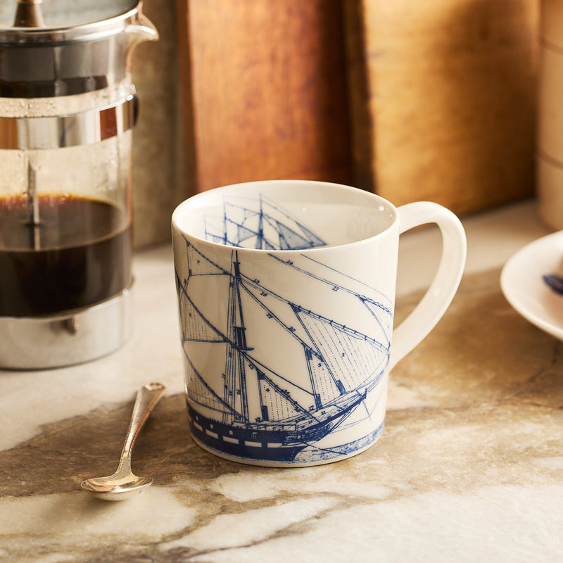 The Caskata Rigging Mug, featuring a blue ship design, rests on a countertop beside a silver spoon and a French press filled with coffee.