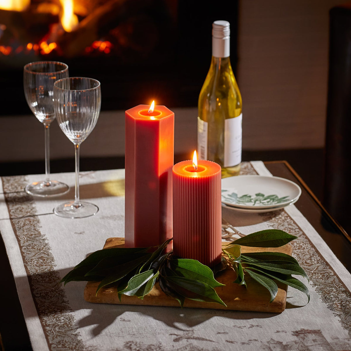 The 9&quot; Rust Hexagon Pillar Candle by Caskata coordinates effortlessly with the ribbed rust candle from the same collection. Here the candles light a romantic table in front of a fireplace.
