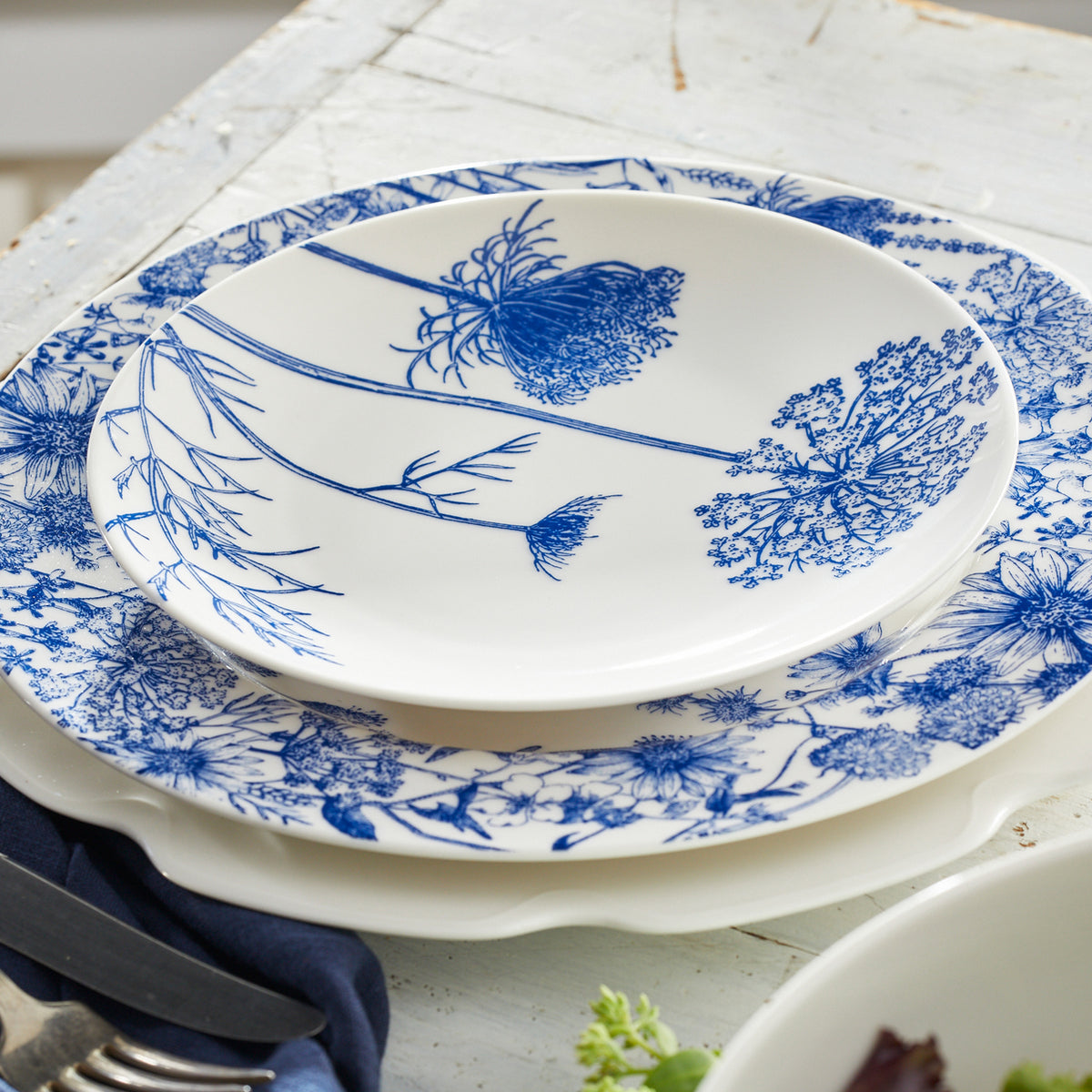 Two white ceramic plates with blue floral patterns from Caskata's Summer Blues Table for 8 set are stacked on a white table, with the top plate showcasing a botanical design; silverware is partially visible beside them.