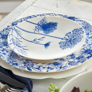 Two white ceramic plates with blue floral patterns from Caskata's Summer Blues Table for 8 set are stacked on a white table, with the top plate showcasing a botanical design; silverware is partially visible beside them.