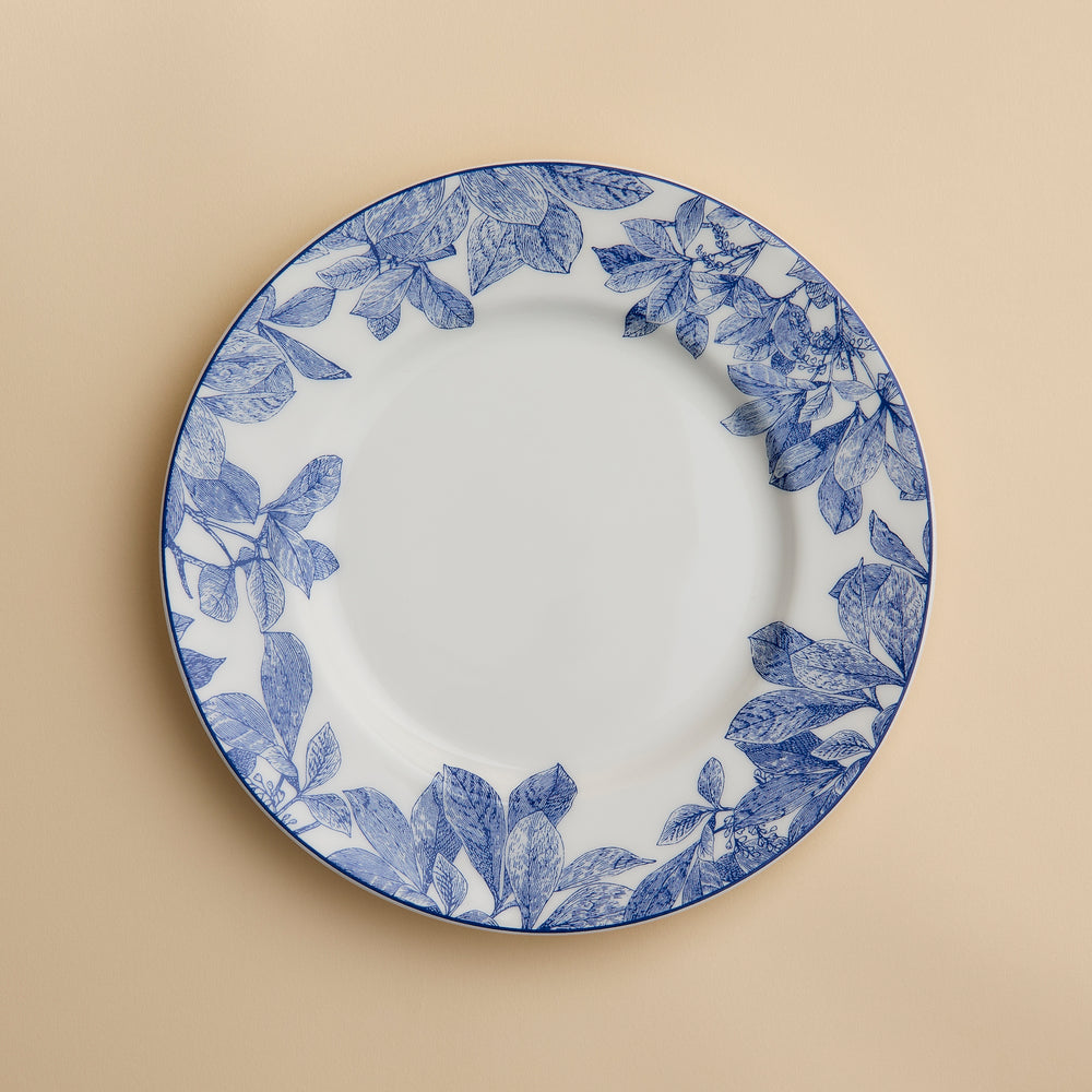 A Caskata Artisanal Home Arbor Rimmed Salad Plate, featuring blue leafy patterns around the white rim, is displayed on a beige background.