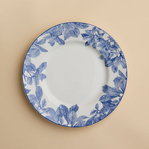 A Caskata Artisanal Home Arbor Rimmed Salad Plate, featuring blue leafy patterns around the white rim, is displayed on a beige background.
