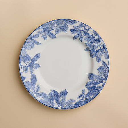 A Caskata Artisanal Home Arbor Rimmed Salad Plate, featuring blue leafy patterns around the white rim, is displayed on a beige background.