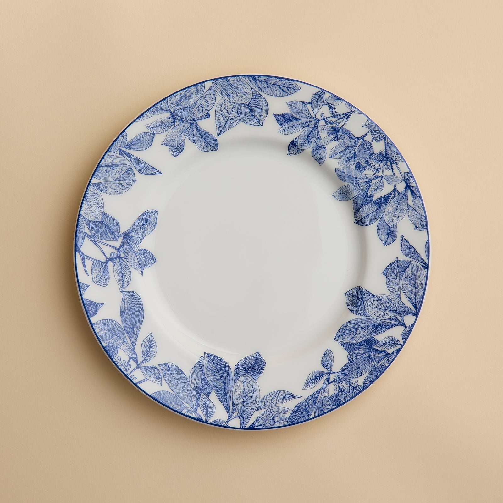 A Caskata Artisanal Home Arbor Rimmed Salad Plate, featuring blue leafy patterns around the white rim, is displayed on a beige background.