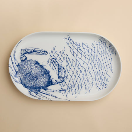 The Caskata Crab Large Coupe Oval Platter showcases a striking blue crab and fishing net illustration on a beige porcelain background, bringing coastal elegance to any table setting.