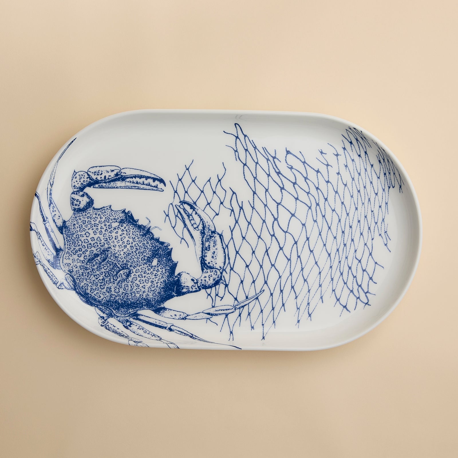 The Caskata Crab Large Coupe Oval Platter showcases a striking blue crab and fishing net illustration on a beige porcelain background, bringing coastal elegance to any table setting.